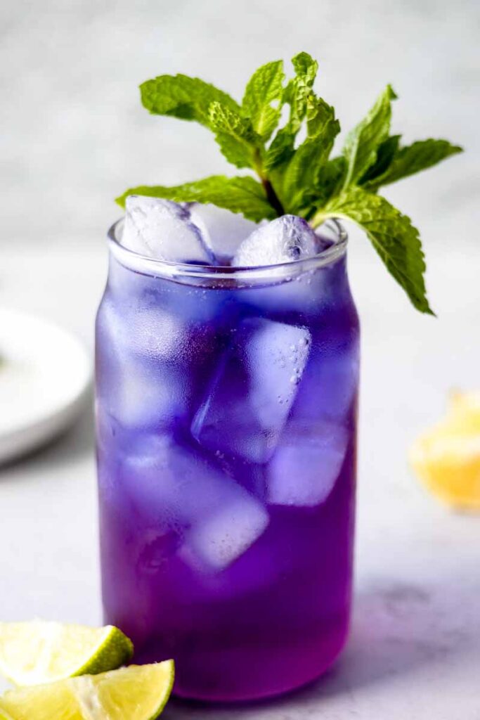 a glass of butterfly pea mocktail with mint garnish and two lime wedges by the side